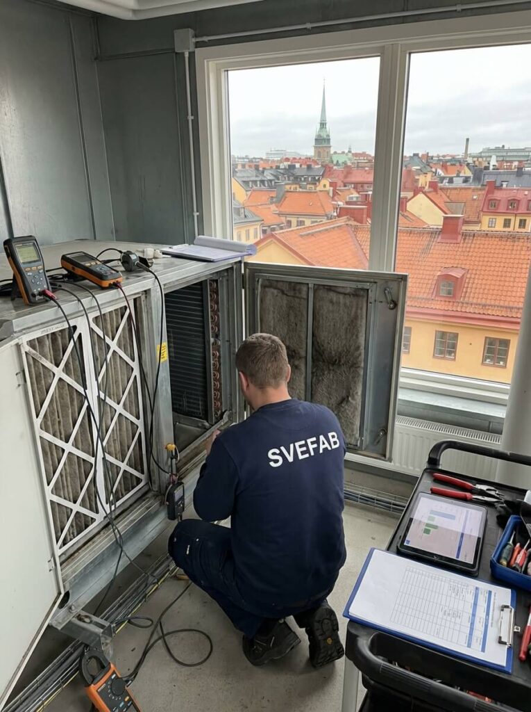 SVEFAB-tekniker i marinblå arbetströja kontrollerar ventilationsaggregat i fläktrum med mätutrustning och Stockholms stadsvy i bakgrunden